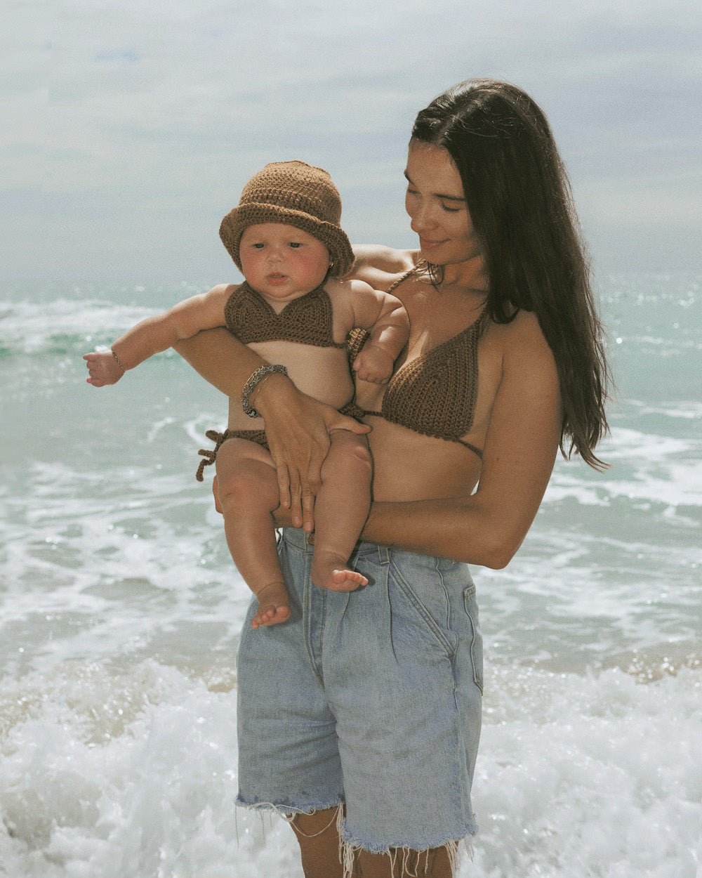 3 MONTH OLD BABY AND MOM IN MATCHING BROWN CROCHET BIKINIS STAND AT THE BEACH